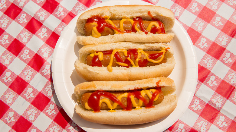 Three hot dogs in buns on paper plate, covered in ketchup and mustard, sitting on checkered tablecloth