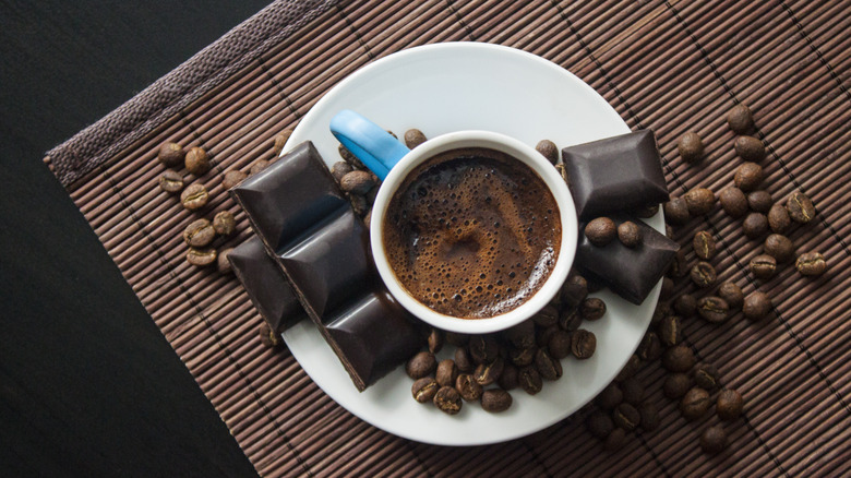 a cup of coffee on small plate next to chocolate bars and coffee beans