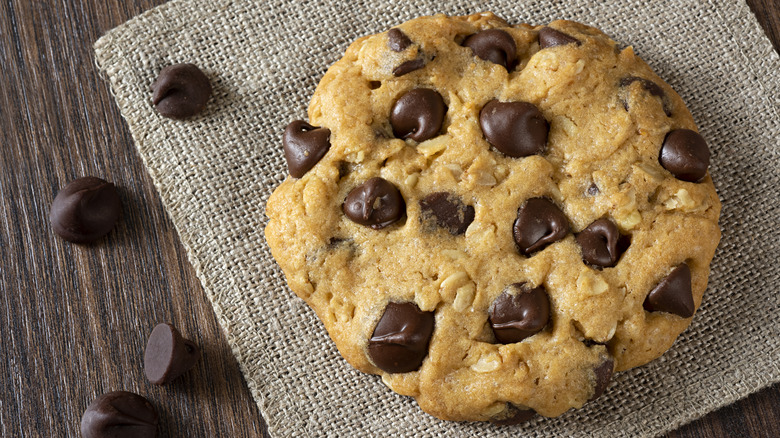 Chocolate chip cookie sitting on woven mat, with chocolate chips next to it