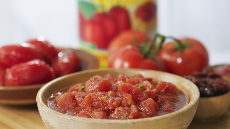 A wooden bowl of diced, herbed tomatoes, with fresh and peeled tomatoes in the background.