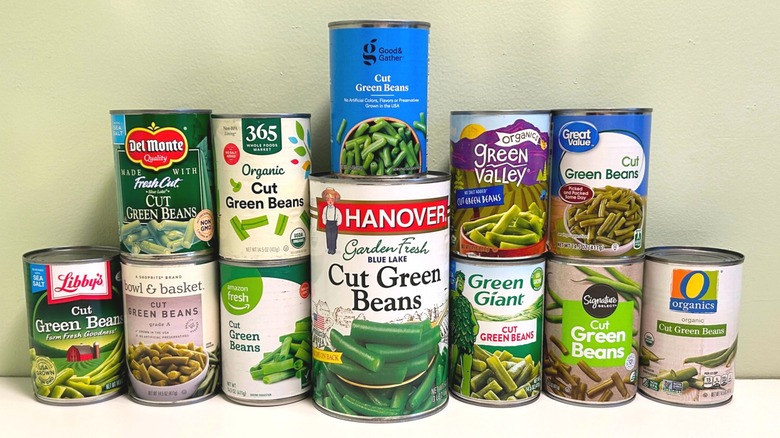 Twelve cans of green beans from different brands lined up on a light background