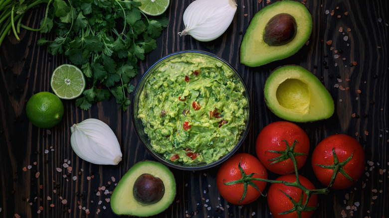 guacamole bowl with limes, onion, tomatoes, and avocados surrounding bowl