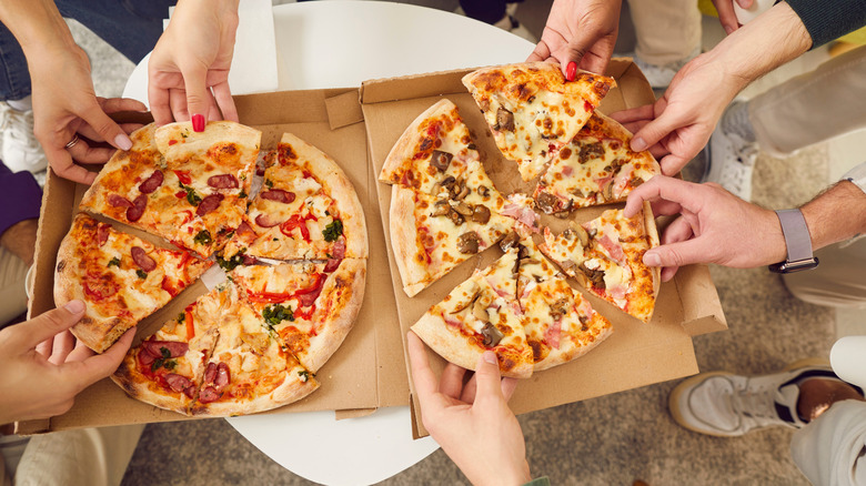 An overhead view of various hands grabbing pizza slices from pizza boxes