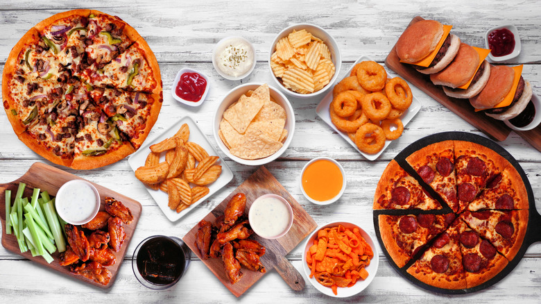 Selection of fast food arranged on table, with pizzas, burgers, chicken wings, onion rings, and various bowls of chips