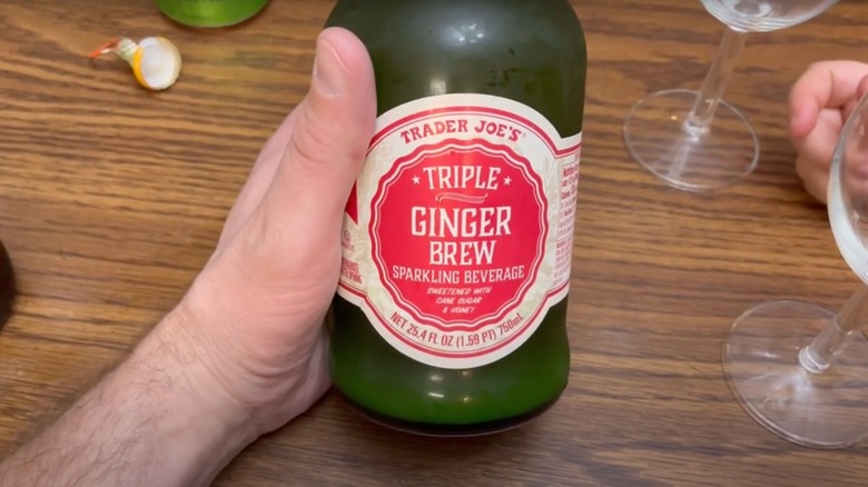 Hand holding a bottle of Trader Joe's Triple Ginger Brew, with wine glasses on table in background