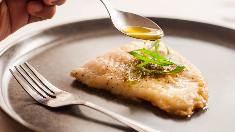 Cooked fish fillet on a gray plate, with butter sauce being spooned over it.