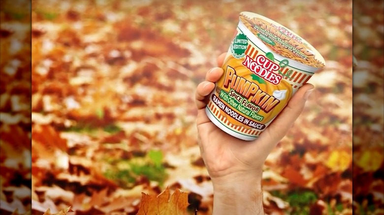 Promotional shot of Pumpkin Spice Cup Noodles, with a hand bursting from autumnal leaves, holding the noodle cup