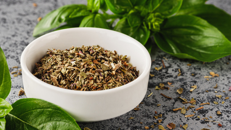 White bowl of dried Italian herbs surrounded by fresh basil leaves