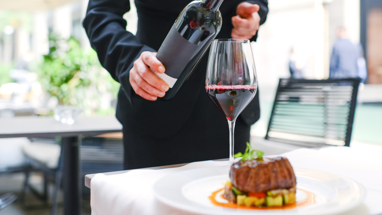 Server pouring wine into glass, next to steak on white dish