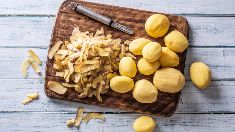 Some potatoes, potato skins, and a peeler placed a cutting board