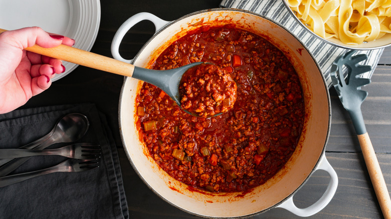 Hand holding a spoonful of bolognese sauce, with pots of sauce and pasta behind it