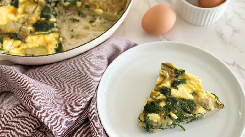 A slice of spinach frittata on a plate next to an egg and a large dish of frittata