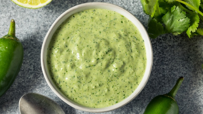 White bowl with aji verde sauce surrounded by cilantro, jalapeño, and lime