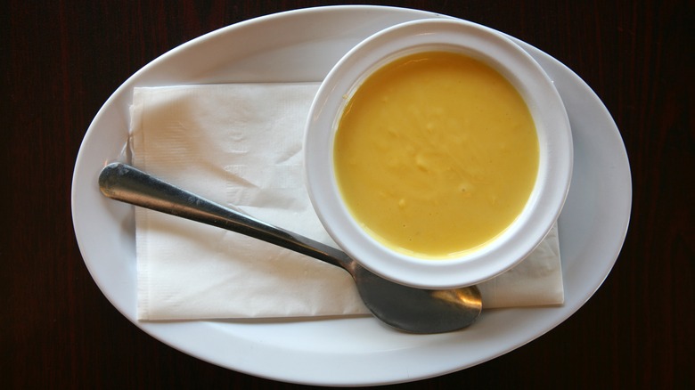 White bowl filled with hollandaise sauce on oval white plate with spoon