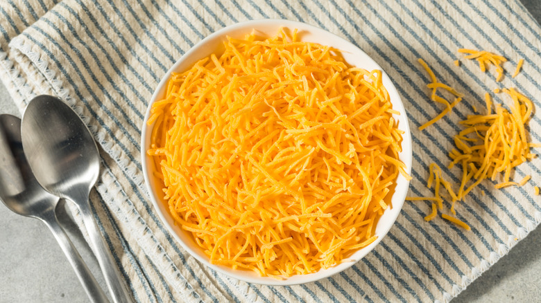 Bowl of finely shredded cheddar cheese on blue-striped napkin with two spoons