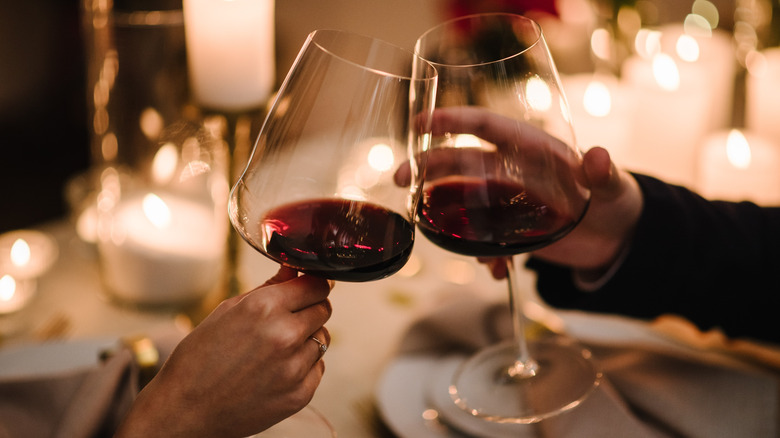 Two red wine glasses being clinked together over a candlelit dinner table.