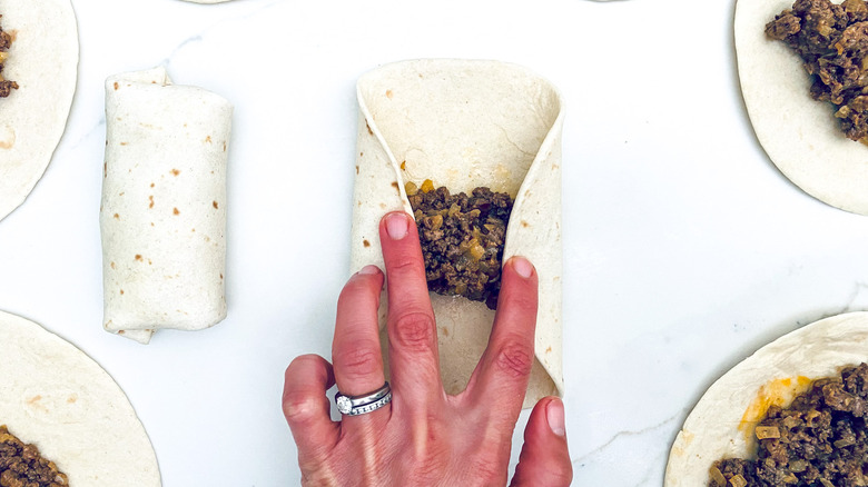 Hand with rings folding tortilla around ground beef