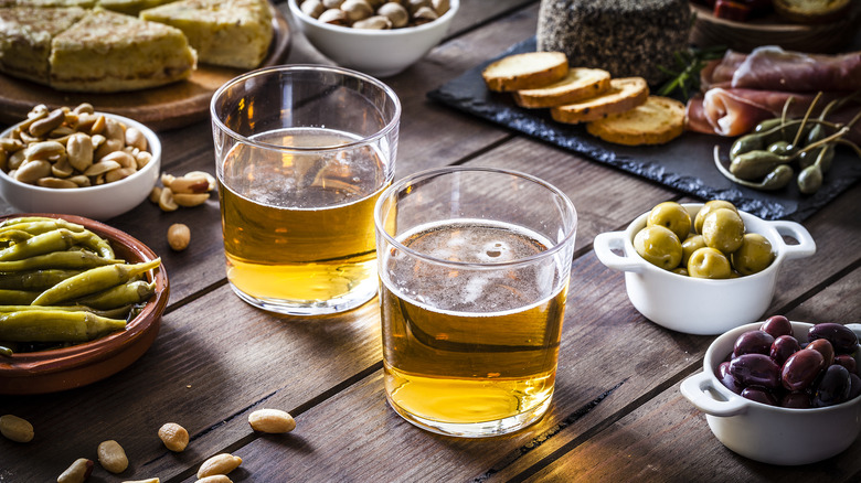 Two short glasses of beer on table with olives and other snacks