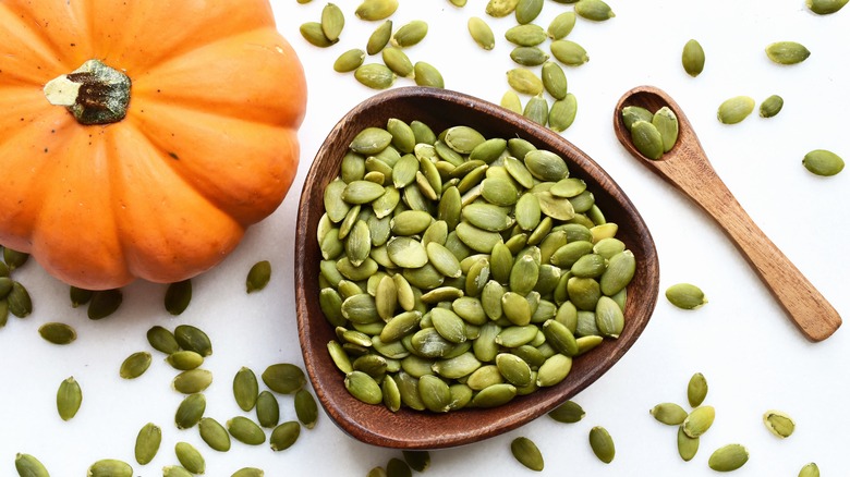 Dish of pumpkin seeds next to a pumpkin on a white surface