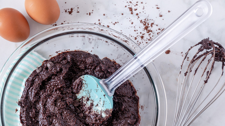 Chocolate chip brownie mix in a bowl with a spatula next to eggs and a hand whisk