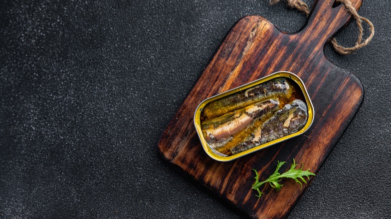 Open can of sardines on wooden cutting board, with a sprig of rocket