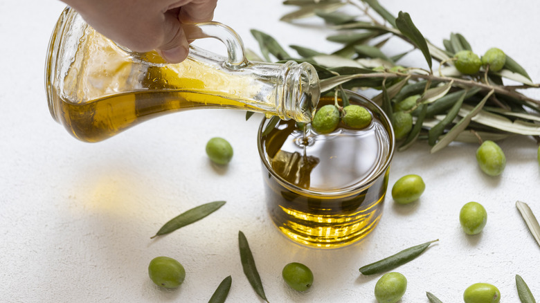 Person pouring olive oil into glass, with olives and olive leaves surrounding it