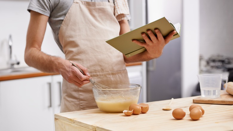 A person whisks batter while holding a cookbook