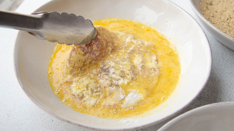 tongs dipping a pork chop in beaten eggs