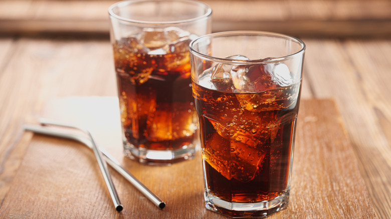 Two glasses of cola with ice on a wooden table beside two metal straws