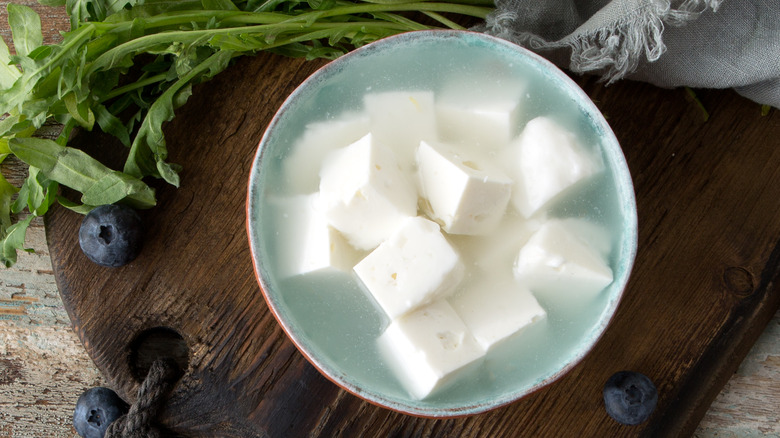 Feta in a bowl of brine, fresh rucola and blueberries on wooden board