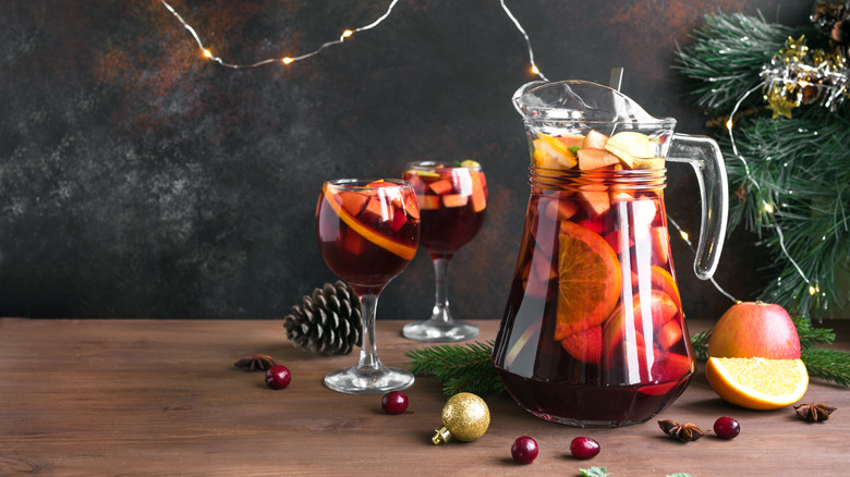 A pitcher of red wine sangria full of cut fruits, with two wine glasses filled to the brim, and a pine tree in the background.