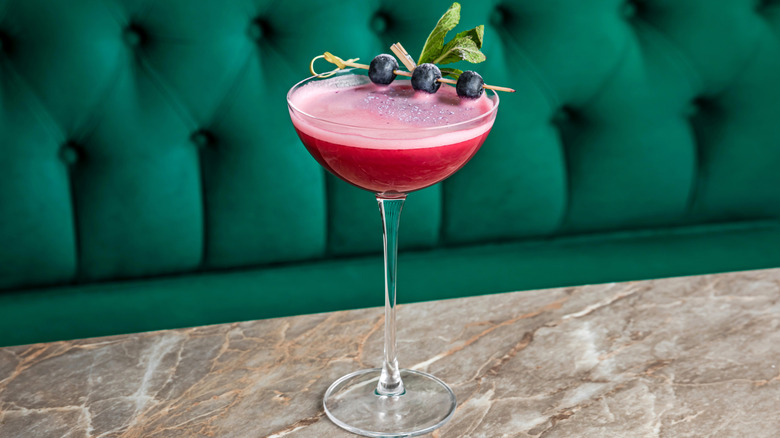 A sour cocktail in a coupe glass garnished with frozen blueberries on a stick