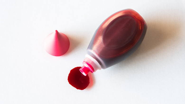 A bottle of red food coloring, cap removed, tipped onto its side to allow some red dye to drip out, forming a blot.