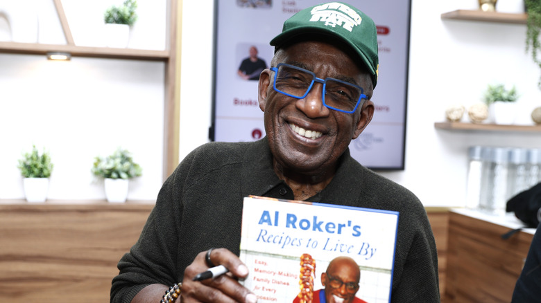 Al Roker wearing blue glasses holding a copy of his cookbook Recipes to Live By