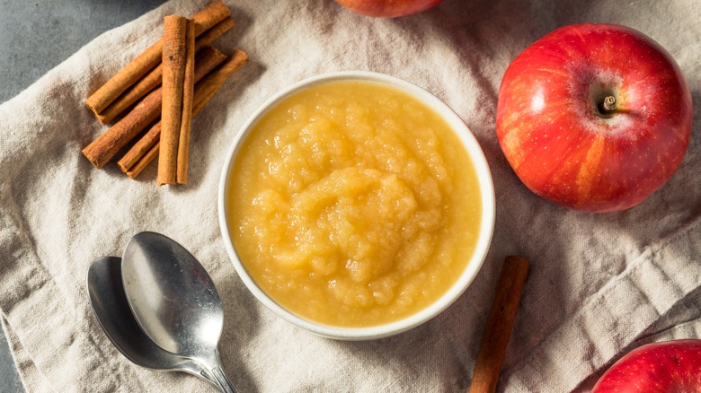 A bowl of applesauce surrounded by a spoon, apples, and cinnamon sticks