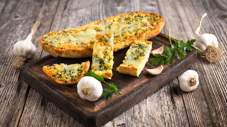 garlic bread on a wood cutting board surrounded by whole garlic