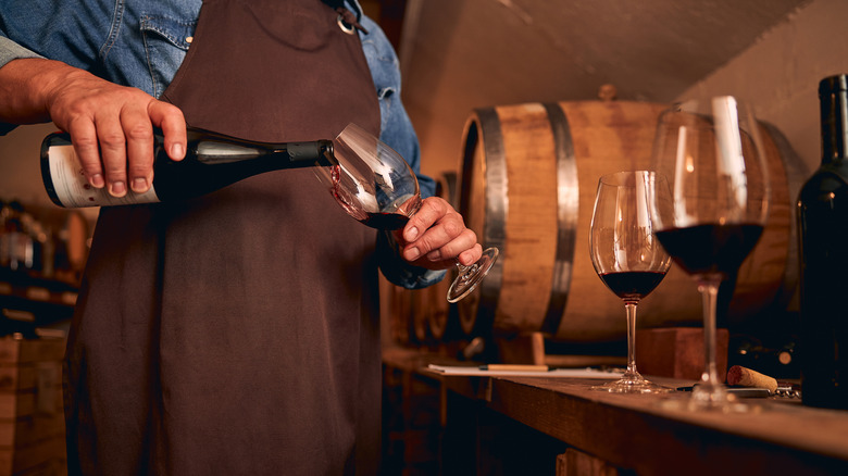 Person pouring red wine into a glass in front of aging barrels