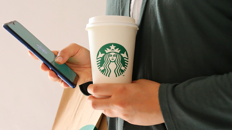 A person holding a Starbucks cup and a telephone with the Starbucks app open.