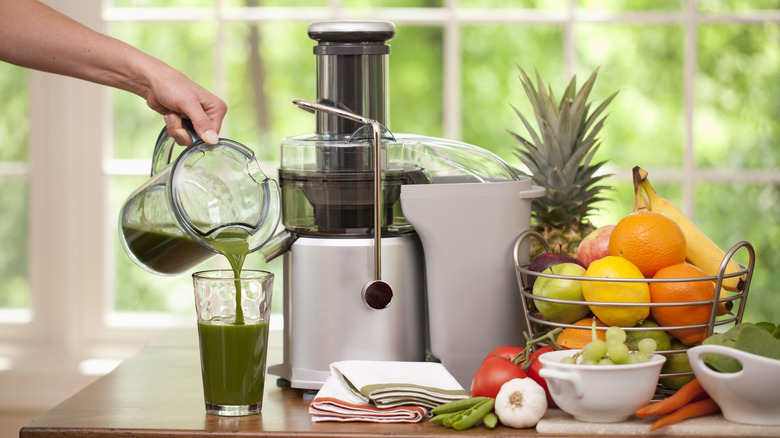 Green juice being poured into a glass by a juicer and bowl of fruit