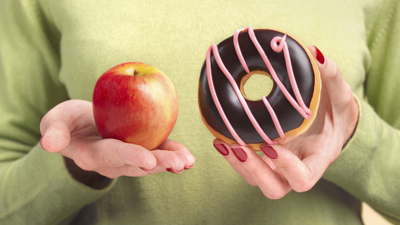 Hands with red-painted nails holds an apple in one and a chocolate-glazed doughnut in the other