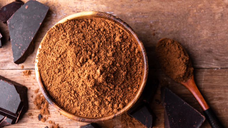 Bowl of cocoa powder on a wood table surrounded with chocolate chunks and a spoon with cocoa powder in it.