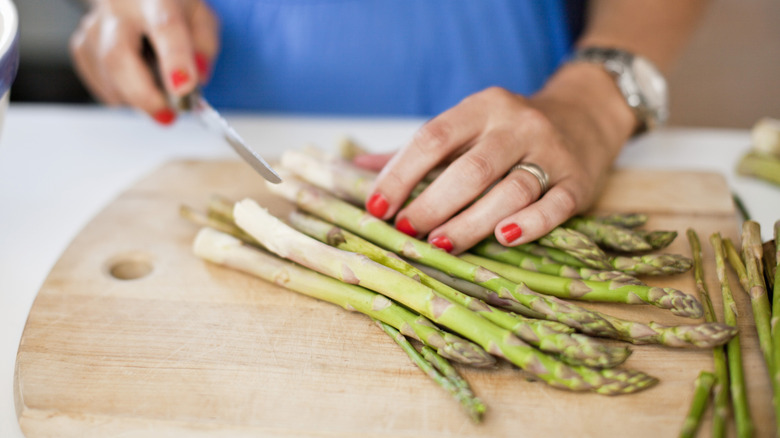 Hands with red nail polish cutting asparagus spears on a wood cutting board