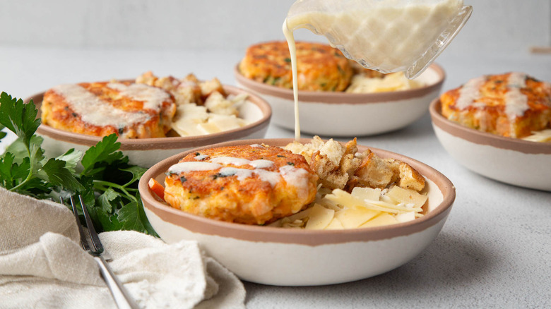 Salad dressing being poured over a salmon burger with coleslaw, croutons, and cheese in a beige bowl