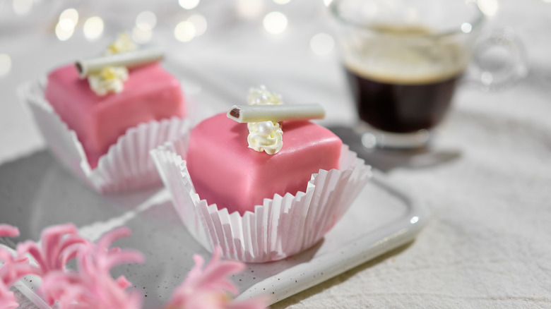 Two pink petit fours on a plate with coffee in background