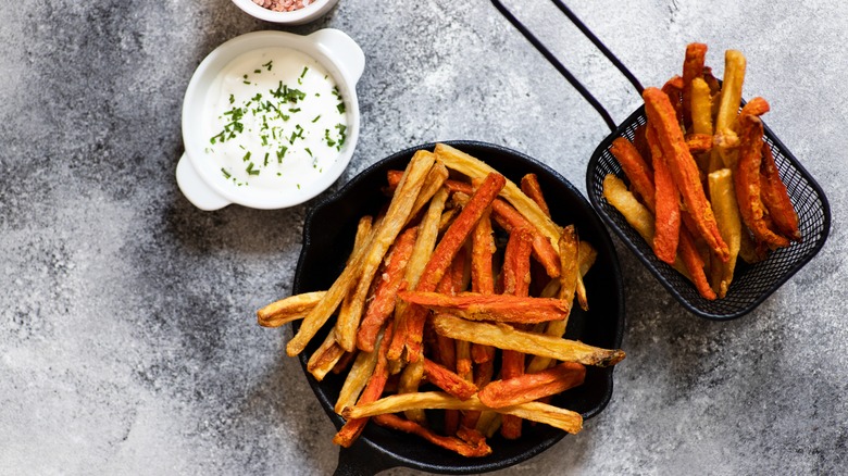 Sweet and white potato fries in a basket and cast-iron skillet with a white sauce in a ramekin