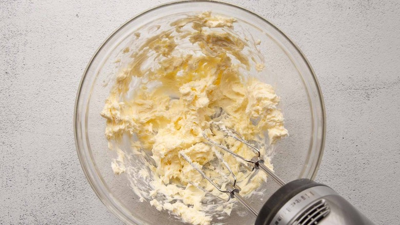 Butter and cream cheese in a clear bowl with a handheld mixer