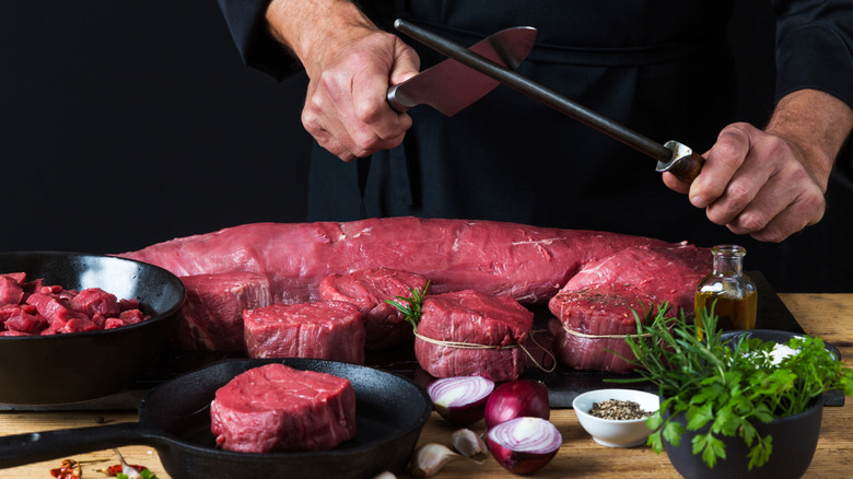 Chef hands honing a knife while cutting meat on counter with herbs and spices