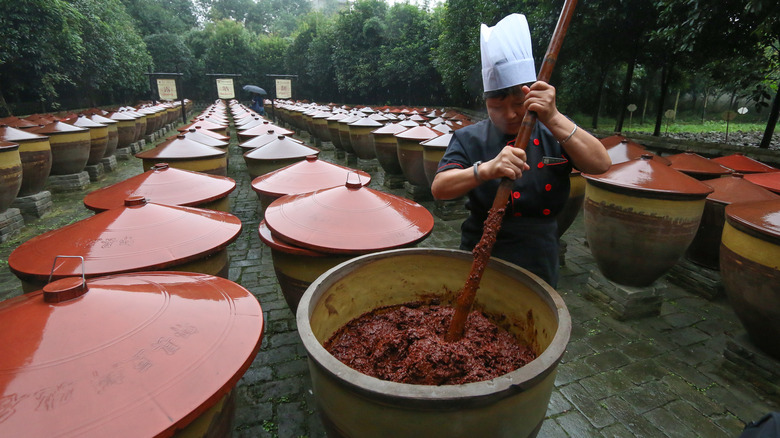 person holding a paddle and mashing dou ban in a large clay vessel