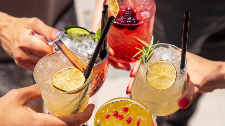 Four hands toasting glasses with colorful cocktails garnishes with fruit.