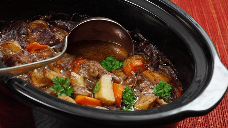 A beef stew with potatoes and carrots with a serving ladle in a black slow cooker.